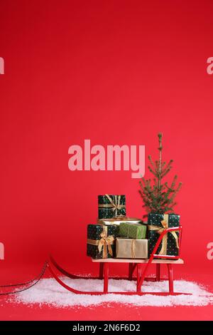 Schlitten mit Geschenkboxen und kleiner Tanne auf rotem Hintergrund. Platz für Text Stockfoto