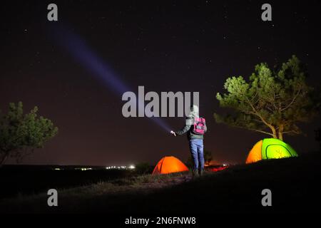 Ein Mann mit heller Taschenlampe in der Nähe von Campingzelten im Freien bei Nacht Stockfoto