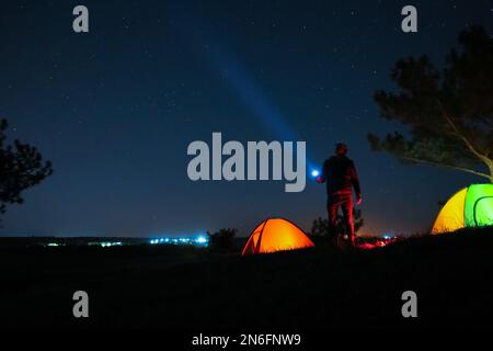 Ein Mann mit heller Taschenlampe in der Nähe von Campingzelten im Freien bei Nacht Stockfoto