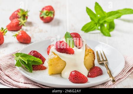 Ein Stück hausgemachter Käsekuchen mit Schlagsahne und frischen Erdbeeren auf weißem Holzhintergrund, dekoriert mit frischen Minzblättern Stockfoto