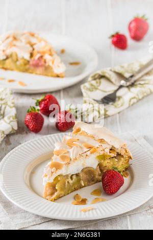 Ein Stück Rhabarberkuchen mit Baiser-Belag, gerösteten Mandeln und frischen Erdbeeren auf weißem Holzhintergrund, vertikal Stockfoto