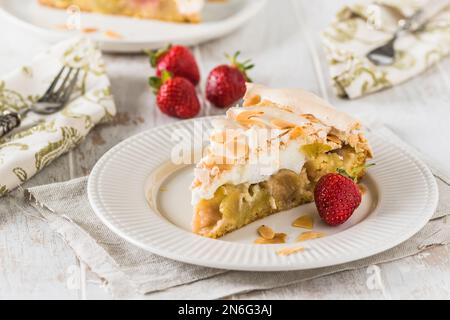 Ein Stück Rhabarberkuchen mit Baiser-Belag, gerösteten Mandeln und frischen Erdbeeren auf weißem Holzhintergrund Stockfoto