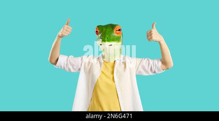 Lustige, humorvolle Frau in der Gummimaske des Froschkopfes zeigt Daumen nach oben auf hellblauem Hintergrund. Stockfoto