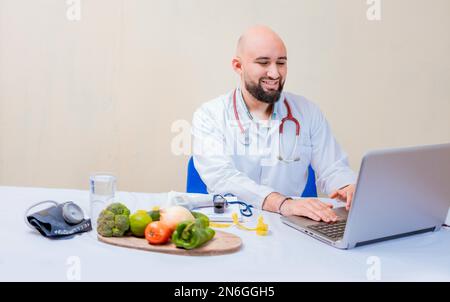 Ernährungsarzt mit Laptop am Arbeitsplatz. Bärtiger Ernährungsberater, der am Schreibtisch an einem Laptop arbeitet, lächelnder Ernährungsberater mit einem Laptop am Schreibtisch Stockfoto