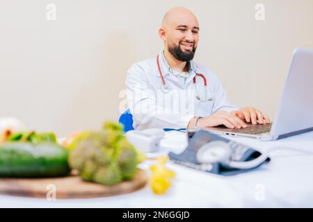 Lächelnder Ernährungsberater mit Laptop am Schreibtisch. Ernährungsarzt mit Laptop am Arbeitsplatz. Ein bärtiger Ernährungsberater, der am Schreibtisch an einem Laptop arbeitet Stockfoto