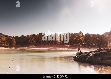 Eine Angelrute steht am Ufer des Flusses vor dem Hintergrund der Silhouetten sprechender Fischer, eines Mädchens und eines Mannes auf den Felsen. Stockfoto