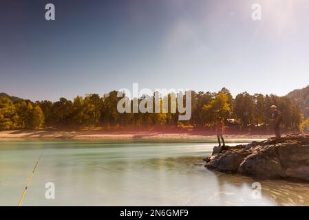 Am Ufer des Flusses steht eine Angelrute vor dem Hintergrund von Silhouetten sprechender Fischer, eines Mädchens und eines Mannes, auf Steinen während des Tages. Stockfoto