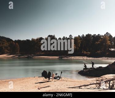 Angelausrüstung steht am sandigen Ufer des Flusses vor dem Hintergrund der Silhouetten von Fischern, einem Mädchen und einem Mann, auf Steinen während der da Stockfoto