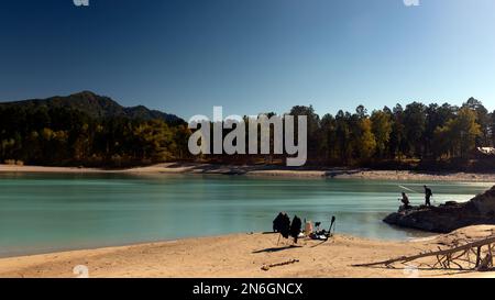 Angelausrüstung steht am Sandufer des Flusses Katun vor dem Hintergrund der Silhouetten von Fischern, einem Mädchen und einem Mann, auf Steinen Stockfoto