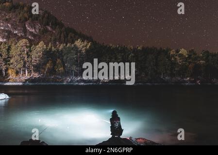 Silhouette eines Fischermädchens, das nachts mit einer Angel und einer Laterne am Flussufer unter einem Berg mit Wald und leuchtenden Sternen sitzt Stockfoto