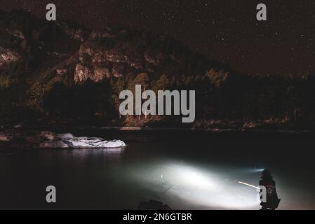 Silhouette eines Fischermädchens, das nachts mit einer Angel und einer Laterne am Flussufer sitzt, unter einem Berg mit Wald und leuchtenden Sternen Stockfoto
