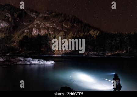 Silhouette eines Fischermädchens, das mit einer Angel und einer Laterne nachts am Flussufer sitzt, unter einem Berg mit Wald und leuchtenden Sternen Stockfoto