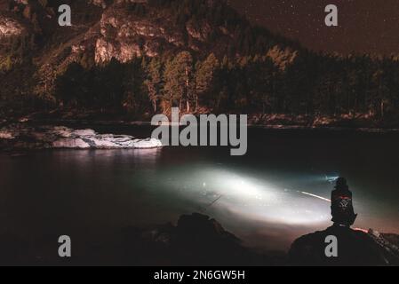 Silhouette eines Fischermädchens, das mit einer Angelrute und einer Laterne bei Nacht am Flussufer sitzt, unter einem Berg mit Wald und hell Stockfoto