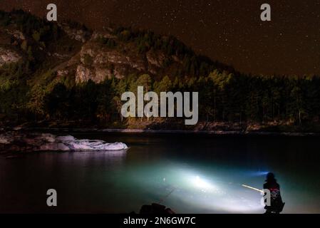 Silhouette eines Fischermädchens, das mit einer Angelrute und einer Laterne bei Nacht am Flussufer sitzt, unter einem Berg mit Wald und hell Stockfoto