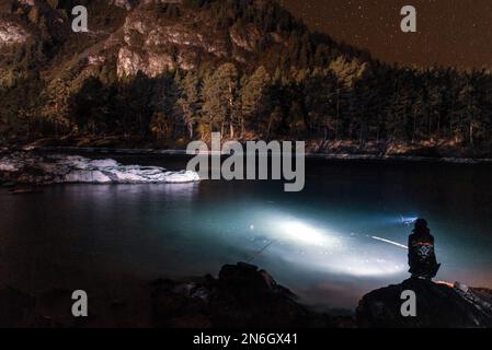 Fischermädchen sitzen mit einer Angelrute und einer Laterne nachts am Flussufer, unter einem Berg mit Wald und leuchtenden Sternen in Altai Stockfoto