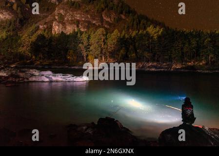 Silhouette eines Fischermädchens, das mit einer Angelrute und einer Laterne bei Nacht am Flussufer sitzt, unter einem Berg mit Bäumen und heller St. Stockfoto