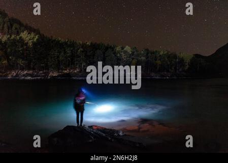 Die Silhouette eines Fischermädchens mit einer Laterne in der Nacht steht im Dunkeln mit einer Angelrute am Flussufer unter einem Berg mit Sternen in Al Stockfoto