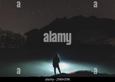 Die Silhouette eines Fischermädchens mit Laterne bei Nacht steht mit einer Angel am Flussufer unter einem Felsen mit Sternen im Altai in Siber Stockfoto