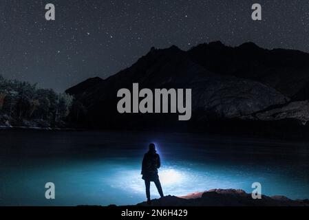 Die Silhouette eines Fischermädchens mit einer Laterne in der Nacht steht im Dunkeln mit einer Angelrute am Flussufer unter einem Felsen mit Sternen in Altai Stockfoto