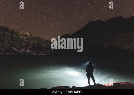 Die Silhouette eines Fischermädchens mit Laterne bei Nacht steht mit einer Angelrute am Flussufer unter einem Felsen mit Sternen im Altai in Sibirien Stockfoto