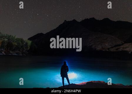 Die Silhouette eines Fischermädchens mit einer Laterne in der Nacht steht im Dunkeln mit einer Angelrute am Flussufer unter einem Felsen mit Sternen in Altai Stockfoto