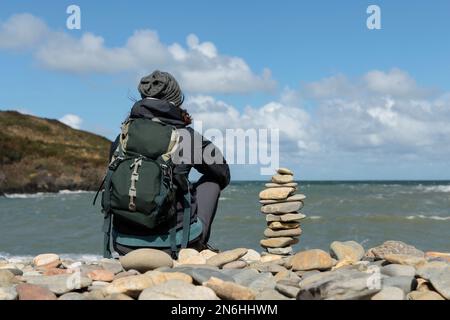 Eine Frau, die allein sitzt und auf das Meer an einem Kieselstrand in Wales schaut. Stockfoto