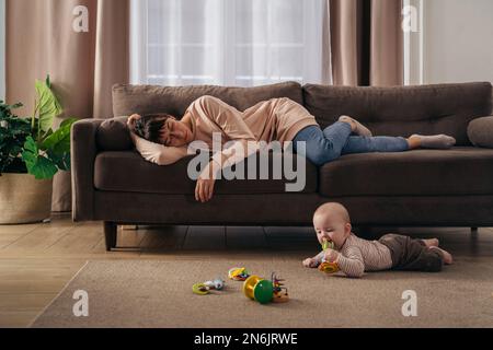 Junge, müde Mutter, die unter Schlafmangel leidet, schläft auf dem Sofa, während ihr kleines Baby auf dem Boden spielt. erschöpfte mutter mit postnataler Depression, will nicht mit ihrem Sohn spielen Stockfoto