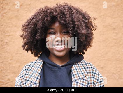 Schwarze Frau, Porträt und Afro an der Wand in der Stadt, in der Stadt oder in Kenia. Glückliche junge afrikanische Frau lächelt für Glück, Gesicht und Locken Stockfoto