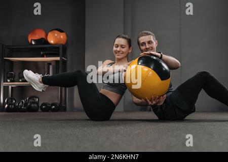 Gesundes junges Paar, das bauchmuskelübungen mit Medizinball im Dark Gym macht. Das Paar lehnt sich Rücken an Rücken auf dem Boden an und trainiert, indem es den Ball an den anderen weitergibt. Sportliche Frauen und Männer trainieren zusammen Stockfoto