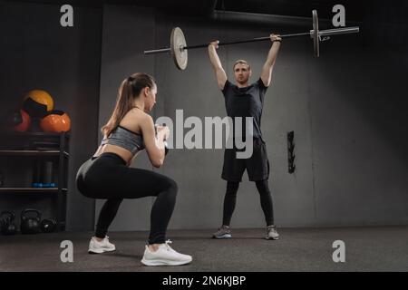 Ein weißes Paar, das Crossfit-Übungen im Dark Gym macht. Attraktive sportliche Frau, die mit Kugelhantel Kniebeugen macht, während ein muskulöser Mann mit schwerer Hantel trainiert. Mann und Frau trainieren zusammen Stockfoto