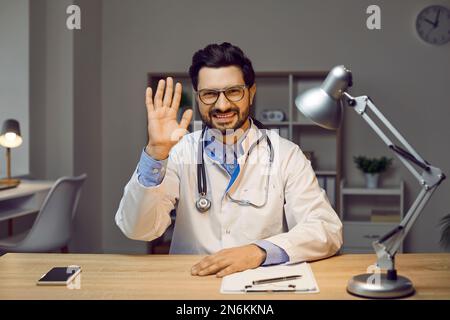 Porträt eines fröhlichen männlichen Arztes, der mit ihm am Tisch sitzt und mit der Hand winkt Stockfoto