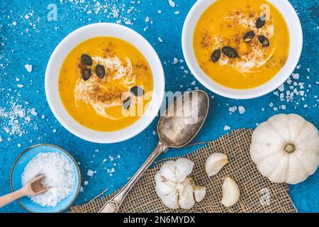 Kürbis-/Karottensuppe in zwei weißen Schüsseln mit Sahne und Kürbiskernen auf blauem Hintergrund. Draufsicht. Stockfoto