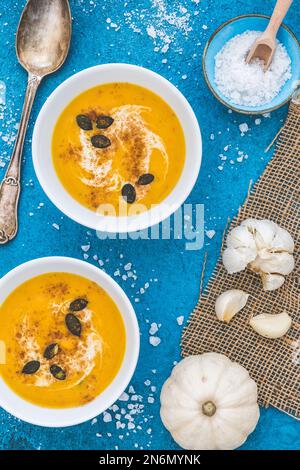 Kürbis-/Karottensuppe in zwei weißen Schüsseln mit Creme und Kürbiskernen auf blauem Hintergrund. Vertikale Ansicht von oben. Stockfoto