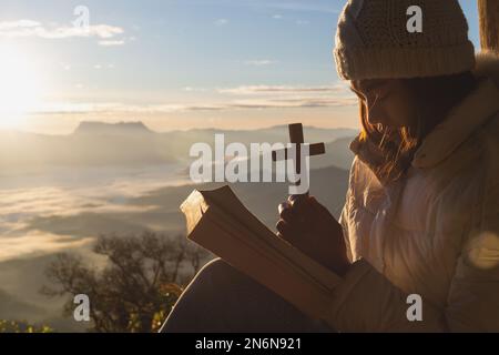 Hände halten ein hölzernes Kreuz über der offenen heiligen bibel auf dem Berghintergrund mit Morgenaufgang, Spiritualität und Religion, Gottesdienst, Sünden und Gebeten. Stockfoto
