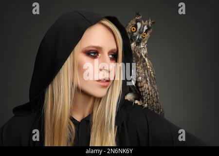 Hexe in schwarzem Mantel mit Eule auf dunklem Hintergrund. Unheimliche Fantasie-Figur Stockfoto