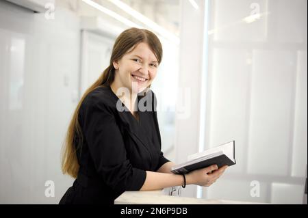 Bild einer jungen Frau, einer Arbeiterin, die lächelt und ein Notizbuch hält, das gegen eine junge Frau steht Bild einer Arbeiterin, die lächelt und ein Notizbuch hält, das vor weißem Hintergrund steht Hochwertiges Foto Stockfoto