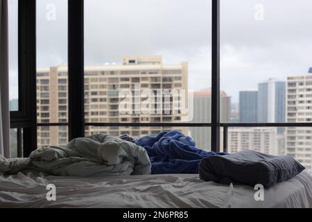 Ungemachtes Bett mit unordentlichen Decken und Kissen in einem Hochhaus-Apartment mit Blick auf die Stadt Stockfoto