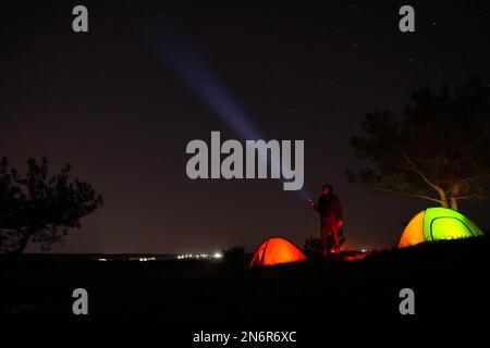 Ein Mann mit heller Taschenlampe in der Nähe von Campingzelten im Freien bei Nacht Stockfoto