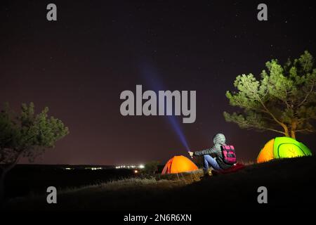 Ein Mann mit heller Taschenlampe in der Nähe von Campingzelten im Freien bei Nacht Stockfoto