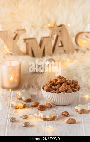 Schüssel mit süßen kandierten Mandeln und Zimtkeksen auf weißem Holz mit Weihnachtsdekoration und Feenlichtern, vertikal Stockfoto