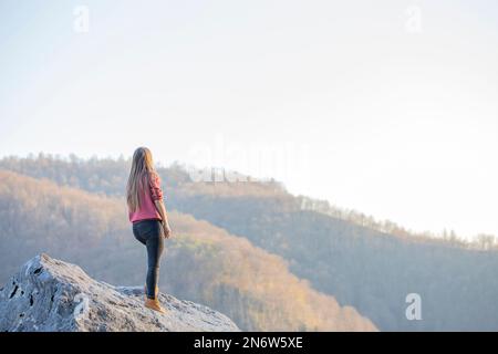 Eine junge Frau genießt die Aussicht vom Gipfel des Berges. Panoramablick auf Berghänge und Wälder im Sonnenlicht Stockfoto