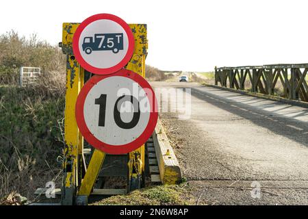 Oberer Fokus eines Gewichts- und Geschwindigkeitsbegrenzungsschilds auf einer temporären Brücke. Ein Auto ist in der Ferne zu sehen, kurz davor, die zerbrechliche Brücke zu überqueren. Stockfoto