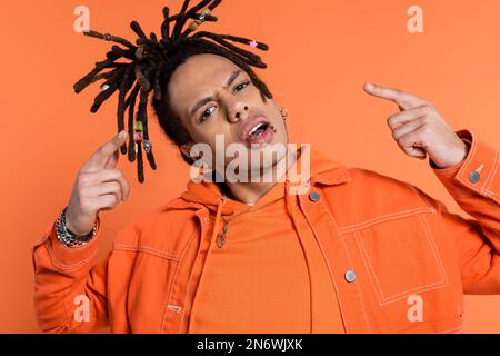 Ein durchbohrter, multiethnischer Mann mit Dreadlocks, der in die Kamera schaut, während er mit isolierten Fingern auf Korallenhintergrund zeigt Stockfoto