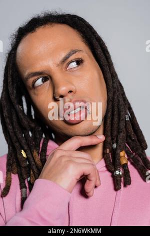Porträt eines pensiven Multirassenmannes in rosa Kapuzenpulli, der isoliert auf grau wegschaut Stockfoto
