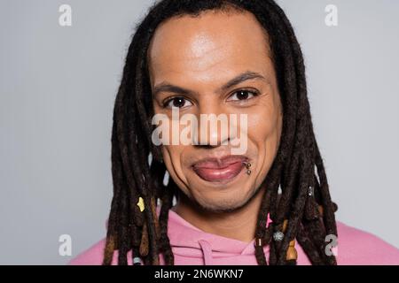 Porträt eines fröhlichen Multirassenmannes in rosa Kapuzenpulli, der auf grau isoliert in die Kamera schaut Stockfoto