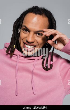 Porträt eines glücklichen Multirassenmannes in rosa Kapuzenpullover, der auf grau isoliert in die Kamera schaut Stockfoto