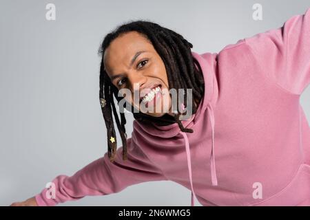 Porträt eines lächelnden Multirassenmannes in rosa Kapuzenpulli, der auf grau isoliert in die Kamera schaut Stockfoto