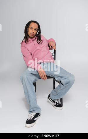 Ein Mann mit mehreren Rassen und durchbohrten Tieren, mit Dreadlocks, die auf die Kamera schauen, während sie auf Grau sitzen Stockfoto