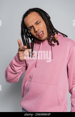 Ein durchbohrter, multiethnischer Mann mit Dreadlocks, der in die Kamera schaut und mit dem Finger auf Grau zeigt Stockfoto