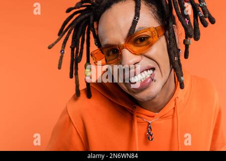 Porträt eines zufriedenen Multirassenmannes mit Kapuzenpullover und stilvoller Sonnenbrille isoliert auf Korallen Stockfoto
